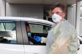 Community nurse wearing a mask and gown as protective wear prepares to a male patient sitting in the car