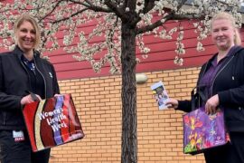 Two female community health nurses standing under a tree with blossoms. They are both holding a women's health week bag and one is holding up information on women's health.