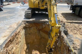 There is a large hole in the road. A yellow digger is removing the earth as part of works to upgrade a sewer main.