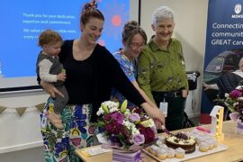Three smiling women stand behind a table. The are cutting a cake that sits on the table together. One of the women is holding a baby. There are flowers and a large light for the one year anniversary celebration.