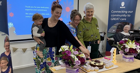 Three smiling women stand behind a table. The are cutting a cake that sits on the table together. One of the women is holding a baby. There are flowers and a large light for the one year anniversary celebration.