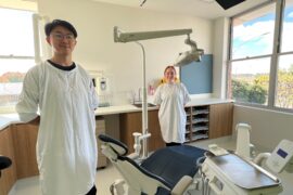A male dentist and female dentist assistant stand next to the dental chair in a new treatment room. They are wearing white gowns and smiling
