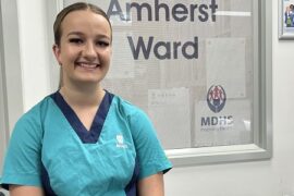 A smiling student nurse stands in front of a window in a hospital ward. She is wearing aqua scrubs.