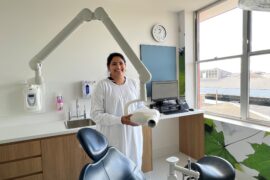 A female dentist is smiling in a new treatment room. She is standing behind the dental chair and holding the x-ray machine