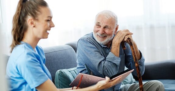 A female home support worker sits with an older man on a catch. They are smiling as the look at a photo album. The man is holding a walking stick.