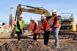 Four people standing on a construction site wearing hard hats and high visibility vests. They are standing in front of a digger and holding shovels loaded with dirt for a sod turn