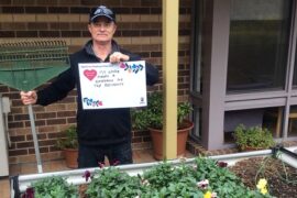 A male male gardener stands behind a raised garden bed at an aged care home. He is wearing a cap and holding a rake in one hand. He holds sign that says 'My work makes a difference for the residents,'
