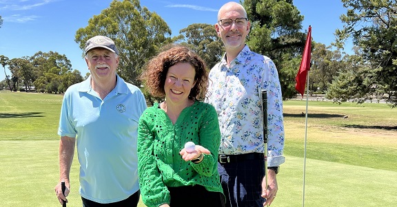 Three people stand on a green on a golf course. They each hold a golf club and there is a flag marking the hole behind them. A women stands in front of men, and reveals a golf ball in the palm of her hand.