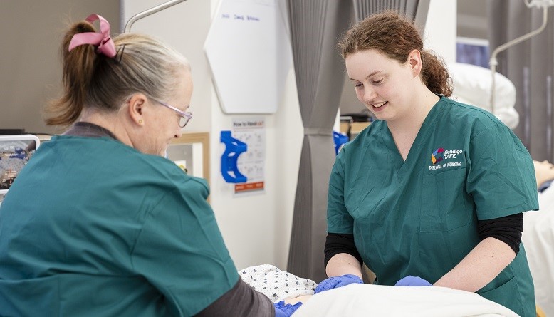 Two student nurses from Bendigo TAFE learn practical skills on a mannequin in a hospital simulation lab.