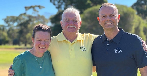 A close up image of three people in a warm embrace. They are wearing casual gear and standing in front of a golf fairway.
