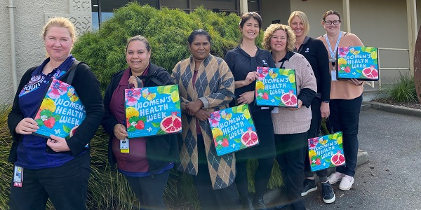 Seven female health care workers stand in front of a hospital smiling. They are holding up colourful tote bags. The text on the bag reads Women's Health Week.