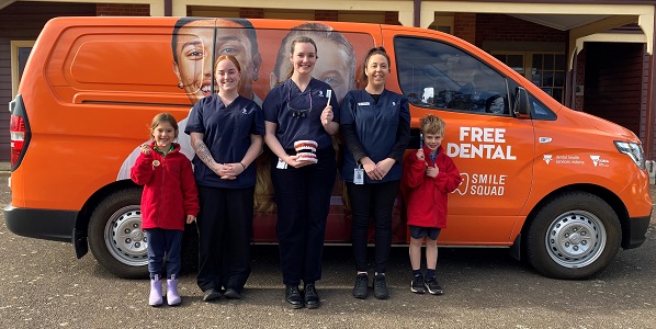 Three young women and two primary school children are standing in front of an orange van. The children are holding a toothbrush in their hand.