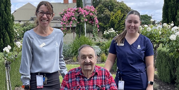 Two member stand next to a male aged care resident in a chair,. The man is sitting in a wheel chair in the garden.