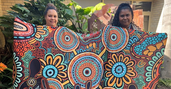 Two First Nations women stand in a gardent., holding up a shawl with an Aboriginal artwork design.