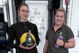 Two female MDHS exercise physiologists are standing before a cable machine with gym equipment, holding dumbells and an exercise ball.