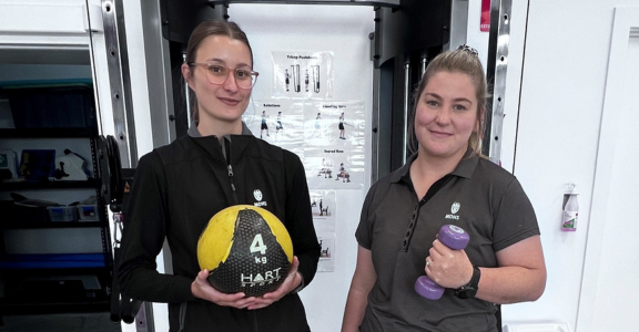 Two female MDHS exercise physiologists are standing before a cable machine with gym equipment, holding dumbells and an exercise ball.