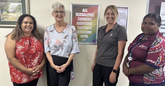 Four female health professionals stand in a row, in front of a gender inclusive sign.