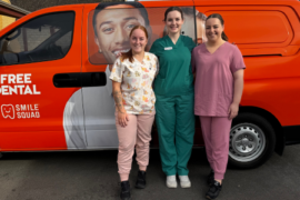 three female MDHS dental services team members in colorful scrubs standing before orange Smile Squad dental van