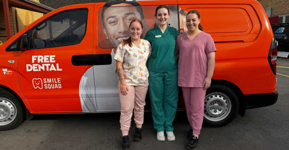 three female MDHS dental services team members in colorful scrubs standing before orange Smile Squad dental van