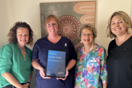 A nurse graduate wearing scrubs holds a certificate. She is standing in a row with female healthcare representatives in casual clothes.