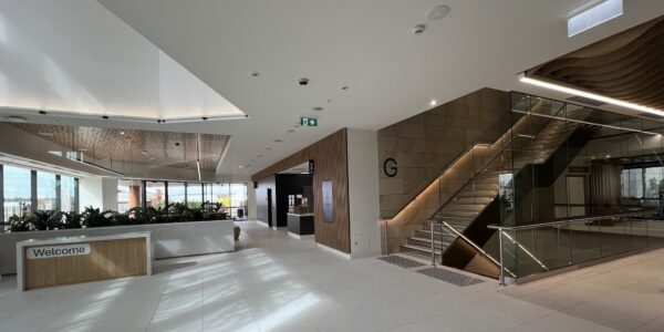 The foyer on the ground level of a new hospital. There is a welcome desk, cafe and stair case on the right.