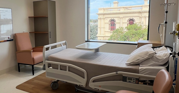 A new of a single room in a new hospital with a bed, beside tray and two chairs. There is a view of a heritage building outside the large window.