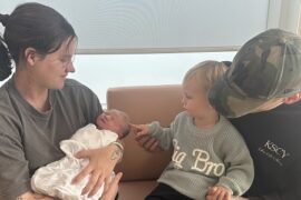 A mum holds her new baby on a couch in a birthing suite. The baby's dad is seated by her side with their young son on his knee.