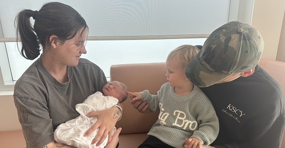 A mum holds her new baby on a couch in a birthing suite. The baby's dad is seated by her side with their young son on his knee.