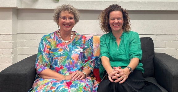 Two female leaders, a CEO and Board Chair, share a moment of connection on a couch. ‘Get involved’ is displayed in white font on an orange box in the top left.”
