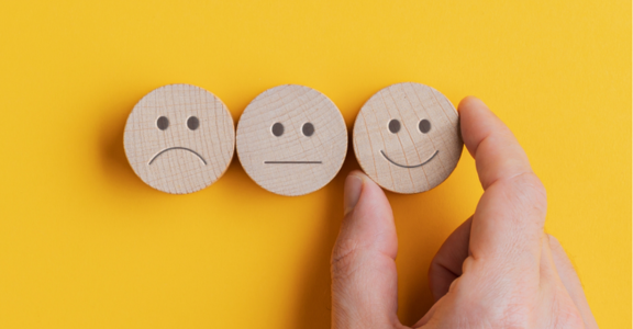 Three wooden emoji tokens sit on a yellow background showing sad, neutral and happy expressions. A hand picks the happy face token.