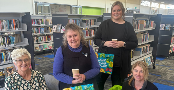 Four people sitting and standing holding tea cups and Women's Health Week Bags