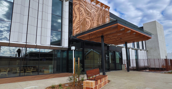 The main entrance to a new hospital. There is a large canopy over the doors and a seat in the forecourt.